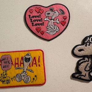 Op de afbeelding: Drie geborduurde patches met Snoopy uit Peanuts. Eén is een roze hart met Snoopy en de woorden "Love! Love! Love!". Een andere is een gele rechthoek met Snoopy en de woorden "HA HA HA!". De derde is Snoopy met een pilotenhelm.