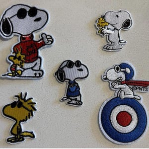 May include: Set of five embroidered patches featuring Snoopy and Woodstock characters. Snoopy is depicted in various poses, including wearing sunglasses, a red sweater, and a pilot's cap. Woodstock is also featured. The patches have a white border.