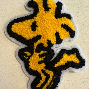 May include: A yellow and black embroidered patch featuring the character Woodstock from the Peanuts comic strip. The patch has a white border and a textured, plush appearance. The design is a cartoon style.