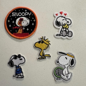 May include: A collection of embroidered patches featuring Snoopy and Woodstock characters. One patch shows Snoopy in a space suit with the word "Snoopy". Other patches depict Snoopy with hearts, sunglasses, and playing tennis. The patches have various colors.