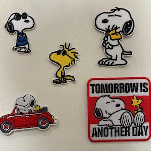 May include: A collection of embroidered patches featuring Snoopy and Woodstock characters. The patches include Snoopy wearing sunglasses, Snoopy in a blue shirt, Woodstock, Snoopy in a red convertible, and a patch with the text "Tomorrow is Another Day."