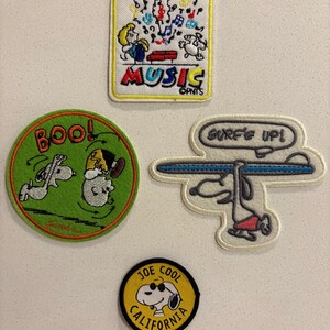 May include: Set of four embroidered patches featuring Snoopy and Woodstock. Designs include "Music" with musical notes, Snoopy surfing with "Surf's Up!", Snoopy and Woodstock with "Boo!", and "Joe Cool California".