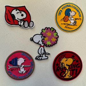 May include: A collection of five embroidered Snoopy patches. One shows Snoopy sleeping in a red doghouse, another with a football, and a third with skis. The others feature Snoopy with flowers and Snoopy with the text "Rocking Out!"
