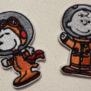 May include: Two embroidered patches featuring Snoopy and Charlie Brown in astronaut suits. Snoopy wears a helmet with goggles, while Charlie Brown has a simple helmet. Both are in orange space suits with black accents and white outlines.