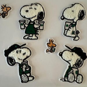 May include: Embroidered patches featuring Snoopy in various poses, some wearing a barista apron and holding coffee cups. The patches are white, black, and green, with a small Woodstock patch included. These patches are ideal for decorating clothing or accessories.