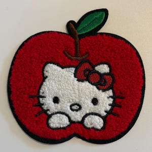 May include: A red apple-shaped patch with a white Hello Kitty design and a red bow. The patch has a black border, a green leaf, and a brown stem. The design is embroidered with a textured, fuzzy appearance.