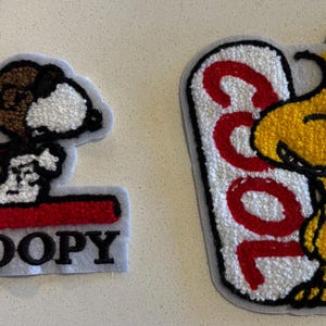 以下が含まれることがあります： ピーナッツのキャラクターが描かれた2つの刺繍パッチ。1つは、名前「SNOOPY」の下にヨガのポーズをとるスヌーピーが描かれています。もう1つは、単語「COOL」のウッドストックが特徴です。どちらのパッチも水色の縁取りがあります。
