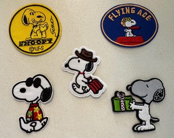 May include: A collection of embroidered Snoopy patches in various designs. One is a yellow circle with Snoopy reading, another is an oval with Snoopy as a Flying Ace, and others show Snoopy in different outfits.