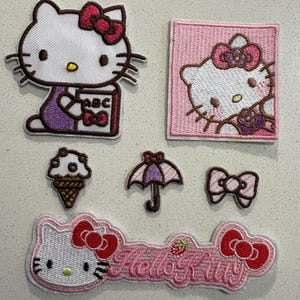 May include: A selection of embroidered Hello Kitty patches. The patches include Hello Kitty holding a book, a pink square patch, an ice cream cone, an umbrella, a bow, and a Hello Kitty banner with the text "Hello Kitty".