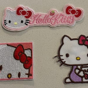 May include: Three embroidered patches featuring the Hello Kitty character. One patch is a rectangular design with the words "Hello Kitty" in pink. The other two patches feature Hello Kitty in different poses, with a red bow.