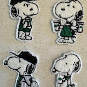 May include: Four embroidered Snoopy patches, each holding a coffee cup. The patches have a white background with black and green detailing. Snoopy is depicted in various poses, wearing a hat or apron. The patches are ideal for clothing.