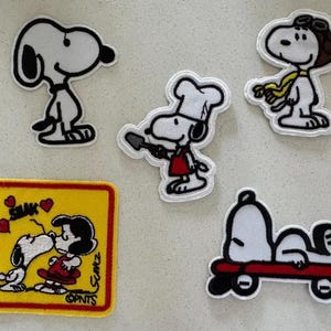 May include: A collection of Snoopy patches in various poses. One patch shows Snoopy as a chef, another as a pilot, and one with Snoopy kissing Lucy. The patches are white with black outlines and accents of red, yellow, and brown.