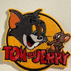 May include: Embroidered patch featuring Tom and Jerry cartoon characters. Tom, a gray cat with orange ears, is depicted with a wide-eyed expression. Jerry, a brown mouse, is also present. The patch has a yellow background with the words "TOM and JERRY" in red.