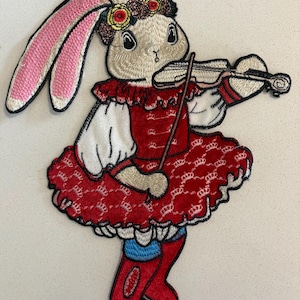 May include: Embroidered patch featuring a bunny playing a violin. The bunny wears a red dress with a crown pattern, a floral crown, and red boots. The patch has a detailed, colorful design with a whimsical, folk art style.