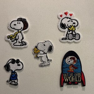 May include: A collection of embroidered Snoopy patches. The patches feature Snoopy in various poses, including an aviator, holding flowers, wearing sunglasses, and in a rocket ship with the text "OUT OF THIS WORLD".