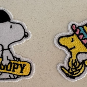 Puede incluir: Dos parches bordados con Snoopy y Woodstock de la tira cómica Peanuts. Snoopy lleva una gorra negra con "NYC" y sostiene una patineta amarilla con "SNOOPY". Woodstock lleva un sombrero a rayas.