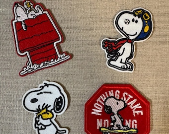 May include: Four embroidered patches featuring Snoopy from the Peanuts comic strip. One patch shows Snoopy on his doghouse, another shows Snoopy as a pilot, a third shows Snoopy holding a yellow bird, and the last one is a red octagon with the text "Nothing Stake No-ing Draw".