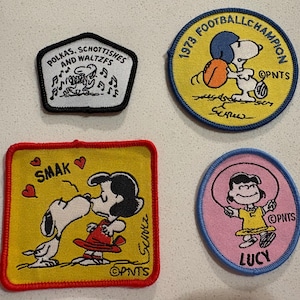 May include: Four embroidered patches featuring Peanuts characters. One patch says "POLKAS, SCHOTTISHES AND WALTZFS". Another shows Snoopy as a football champion with the text "1973 FOOTBALL CHAMPION". A third patch shows Snoopy kissing Lucy with the word "SMAK". The last patch shows Lucy jumping rope with the word "LUCY".
