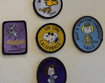 May include: A collection of embroidered Peanuts character patches. The patches feature Snoopy with sunglasses, Woodstock, Sally, Peppermint Patty, and a character saying "AAUGHH!". Each patch has a different color scheme and text.