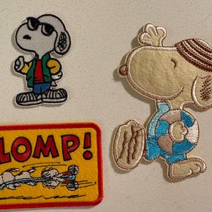 以下が含まれることがあります： スヌーピーのキャラクターが描かれた3つの刺繍パッチ。1つはサングラスとジャケットを着たスヌーピー。もう1つは水着姿のスヌーピー。3つ目は「CLOMP！」の文字とスヌーピーの画像が入った黄色の長方形のパッチです。