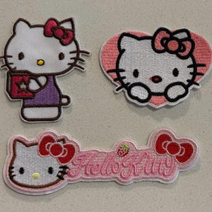 Op de afbeelding: Drie geborduurde Hello Kitty-patches. Eén toont Hello Kitty met een boek, een andere is hartvormig en de derde spelt "Hello Kitty" met een aardbei. Allemaal hebben ze een witte achtergrond met roze en rode accenten. Ideaal voor kleding of accessoires.