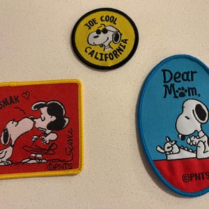 May include: Three embroidered patches featuring Snoopy and related characters. One patch is round and yellow with "JOE COOL CALIFORNIA" text. Another is red and square with Snoopy kissing Lucy. The third is blue and oval with Snoopy and the text "Dear Mom."