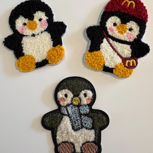 Könnte beinhalten: Drei gestickte Pinguin-Patches. Ein schwarz-weißer Pinguin mit orangefarbenen Füßen und einem gelben Schnabel. Ein weiterer Pinguin trägt eine rote Mütze und eine Handtasche mit goldenem Logo. Der dritte Pinguin hat einen blauen Schal.