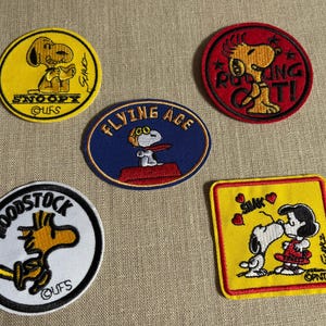 以下が含まれることがあります： ピーナッツのキャラクターが描かれた刺繍ワッペン5枚のコレクション。スヌーピー、ウッドストック、ルーシーなどのデザインがあり、「Flying Ace」や「Rocking It！」などの文字も含まれています。様々な形と色のワッペンです。