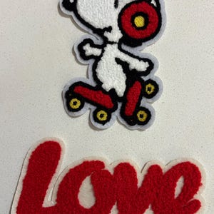 Può includere: Toppe ricamate raffiguranti Snoopy sui pattini a rotelle con ruote e cuffie rosse, e la parola "Love" in rosso, il tutto con bordi bianchi. Le toppe sono su una superficie bianca.