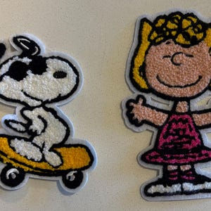 以下が含まれることがあります： 漫画のキャラクターが描かれた2つの刺繍パッチ。1つは、黄色のスケートボードに乗った白と黒の犬です。もう1つは、腕を広げたピンクのドレスを着た女の子です。どちらも白い縁取りがあります。