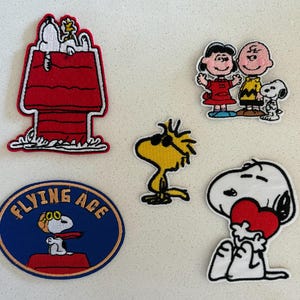May include: A collection of embroidered patches featuring characters from the Peanuts comic strip. Includes Snoopy in his doghouse, Snoopy with a heart, Woodstock, and Charlie Brown with Lucy and Snoopy. The patches have various colors and designs.