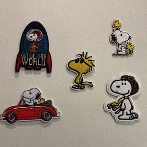 May include: Embroidered patches featuring Snoopy and Woodstock characters. Includes Snoopy in a rocket ship with "OUT OF THIS WORLD" text, Snoopy driving a red car, and Snoopy with a pilot's cap. Also includes Woodstock patches.