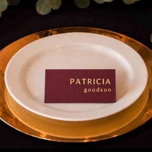 May include: A rectangular burgundy place card with gold lettering reading PATRICIA goodson, centered on a white dinner plate. The plate rests on a large, polished gold charger plate, positioned on a black tablecloth with a gold fork and glass.