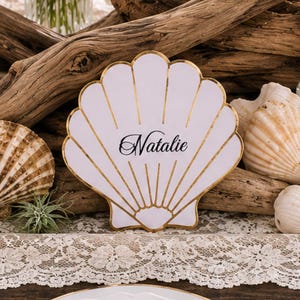 Op de afbeelding: Een witte schelpvormige naamkaart met goudfolie omlijning en de naam Natalie in zwarte schrijfletters. De kaart staat tussen drijfhout, diverse natuurlijke schelpen en glazen flesjes op een tafelkleed met een kanten rand.