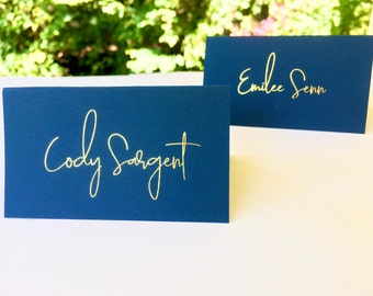 May include: Navy blue place cards with gold calligraphy. One card reads "Cody Sargent" and the other reads "Emilee Senn". The cards are rectangular and stand upright. Ideal for weddings, parties, or special events.
