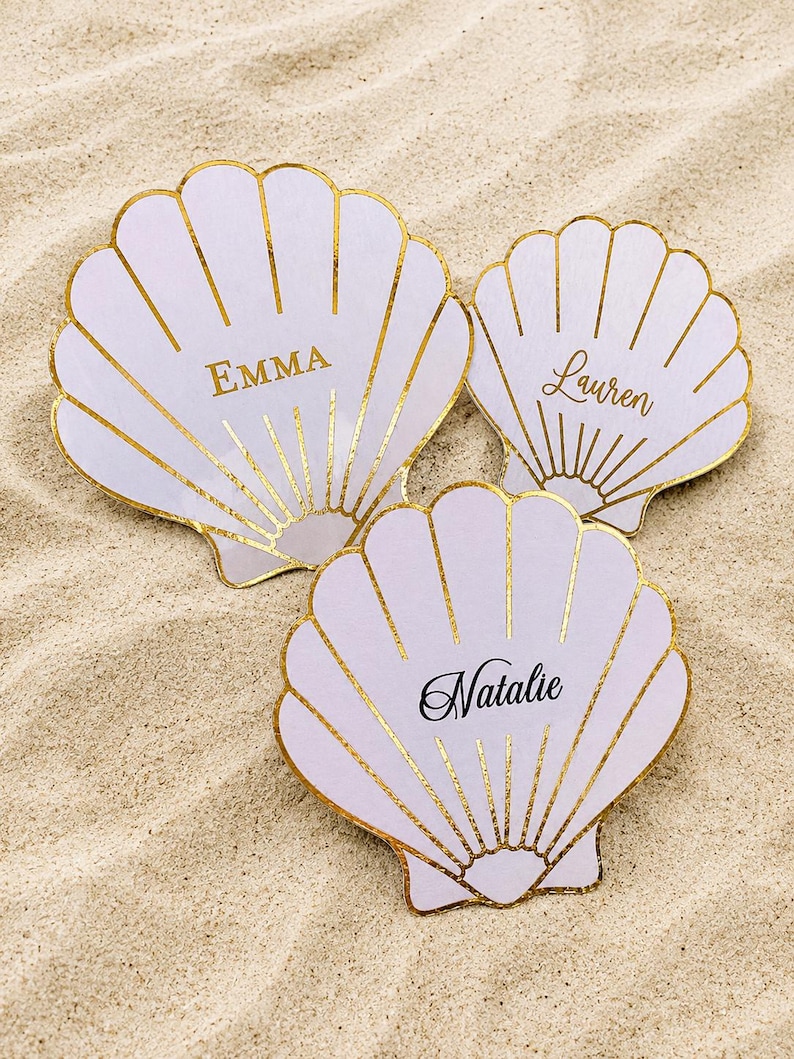 Op de afbeelding: Drie lichtpaarse schelpvormige kaarten met goudfolie omlijningen. Elke schelp is voorzien van een naam in goud of zwart: Emma, Lauren en Natalie. De kaarten hebben een geschulpt randontwerp en zijn gerangschikt op een zandige ondergrond.