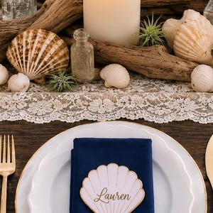 Op de afbeelding: Een wit dinerbord met een gouden rand bevat een gevouwen marineblauw servet, bekroond met een witte schelpvormige naamkaart met de naam Lauren in gouden letters. De tafelsetting bevat een gouden vork en een middelpunt van drijfhout en schelpen.