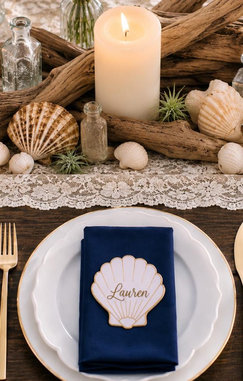 Op de afbeelding: Een wit dinerbord met een gouden rand bevat een gevouwen marineblauw servet, bekroond met een witte schelpvormige naamkaart met de naam Lauren in gouden letters. De tafelsetting bevat een gouden vork en een middelpunt van drijfhout en schelpen.