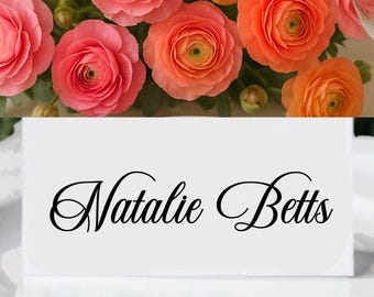 May include: A white place card with the name "Natalie Betts" in elegant black script sits on a white plate. Above, a collection of pink and orange ranunculus flowers adds a vibrant floral touch.