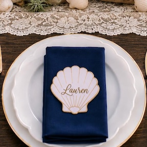 Op de afbeelding: Een wit bord met een geschulpte rand en een gouden bies bevat een gevouwen marineblauwe stoffen servet. In het midden van het servet ligt een wit, schelpvormig naamkaartje met een gouden rand en de naam Lauren in gouden cursief schrift.