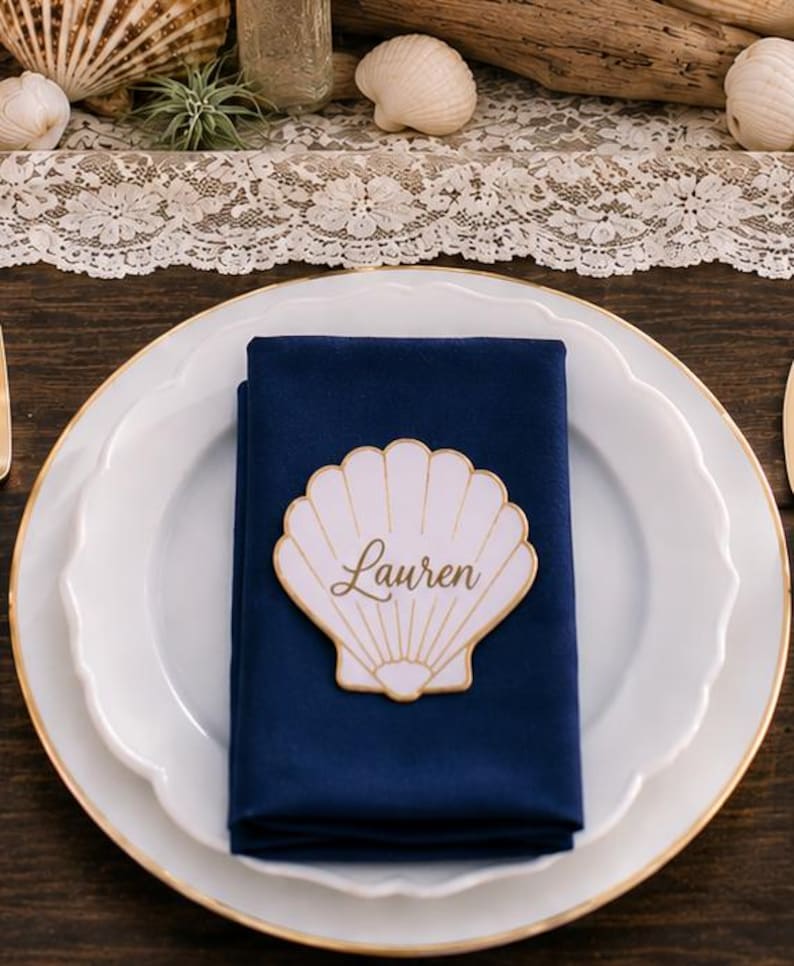 Op de afbeelding: Een wit bord met een geschulpte rand en een gouden bies bevat een gevouwen marineblauwe stoffen servet. In het midden van het servet ligt een wit, schelpvormig naamkaartje met een gouden rand en de naam Lauren in gouden cursief schrift.