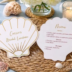 May include: Two white, shell-shaped paper cards with gold foil outlines. The left card reads "Carrie's Bridal Luncheon" in gold script. The right card features the word "Menu" in gold script, followed by a list of starters, desserts, and beverages.