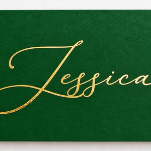 May include: A rectangular dark green card featuring the name Jessica written in a gold cursive script font. The surface of the card has a textured finish, and the gold lettering is centered horizontally and vertically on the green background.