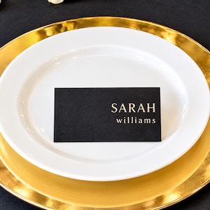 May include: A rectangular black place card with gold foil lettering reading SARAH williams, centered on a white dinner plate which is resting on a large gold charger plate. The place card features a clean, minimalist design with serif font.