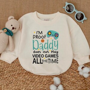 May include: Cream-colored sweatshirt with the text "I'M PROOF Daddy does not play VIDEO GAMES ALL THE TIME" and a video game controller graphic. The sweatshirt also has a small tag that says "Stofaz".