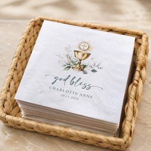 May include: A stack of white paper napkins featuring a gold chalice illustration with a cross, green foliage, and the text "god bless", "CHARLOTTE ANNE", and "20.11.2026" printed in dark green and black ink on the front surface of the top napkin.