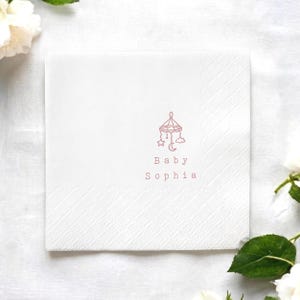 May include: A square white paper napkin featuring a delicate pink line drawing of a baby mobile with a star, moon, and cloud. Below the mobile, the text reads Baby Sophia in a simple, clean font. The napkin has a textured, embossed border.