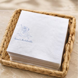 May include: A stack of white paper napkins featuring a blue line art illustration of a person in a sun hat and sunglasses floating on an inner tube while holding a drink, with the text Emma's Bachelorette printed in blue script below the graphic.