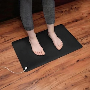 May include: A black rectangular mat with a white cord and plug. The mat is on a wooden floor. The image shows a person standing on the mat.