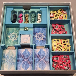 Könnte beinhalten: Ein Brettspiel-Organizer mit verschiedenen Komponenten. Der Organizer hat mehrere Fächer, die mit Spielsteinen in Farben wie Schwarz, Pink, Gold, Grün und Weiß gefüllt sind. Es gibt auch Karten und Spielsteine mit grafischen Designs und dem Wort "FISH".